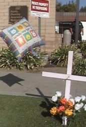 White cross next to driveway of Bakersfield abortion chamber, with flowers and a balloon in memory of the aborted children.